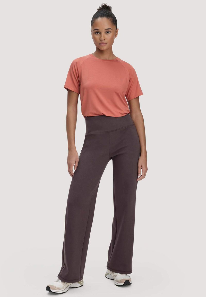 Vrouw staand, gekleed in een roestkleurig T-shirt met korte mouwen, hooggetailleerde donkerbruine brede broekspijpen en lichtgekleurde sneakers.