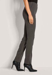 Vrouw in zwarte gebloemde broek en zwarte hoge hakken enkellaarsjes, staande in profiel tegen een lichte achtergrond.