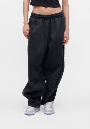 Personne portant un pantalon de survêtement noir ample, des baskets blanches et grises, et un top noir court, debout devant un fond uni.
