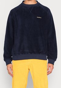Felpa in pile blu navy con collo e polsini a costine, caratterizzata da un piccolo logo dorato "woodbird" sul petto. Indossata con pantaloni gialli in velluto a coste.