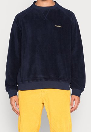 Sudadera de forro polar azul marino con cuello redondo y puños acanalados, que presenta un pequeño logotipo dorado de "woodbird" en el pecho. Combinada con pantalones de pana amarillos.