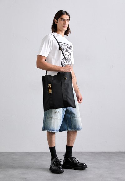 Geantă tote din nylon negru cu logo auriu, având mânere multiple și un bandulator ajustabil. Modelul poartă un tricou cu grafică și șorturi din denim.