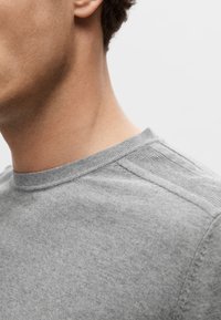 Szary t-shirt z dzianiny z okrągłym dekoltem, subtelną teksturą i pionowymi akcentami w prążki na ramionach. Zbliżenie na tors.