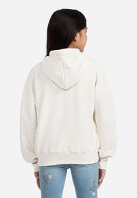 Giovane con lunghi capelli scuri che indossa un felpa crema e jeans azzurri chiari con ricami floreali, di spalle contro uno sfondo sobrio.