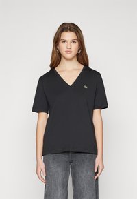 T-shirt nera a V in cotone, con maniche corte e un piccolo logo verde sul lato sinistro del petto. Tessuto liscio, vestibilità rilassata.
