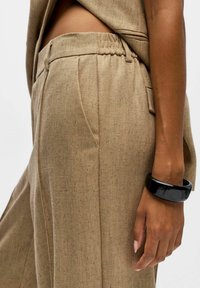 Pantalons beige à motif en chevrons avec une taille élastique et une poche latérale, accompagnés d'un bracelet noir brillant au poignet.