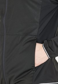 Zwarte jas van gladde stof, met een voorrits, pocketdetails en witte accenten op de manchetten.