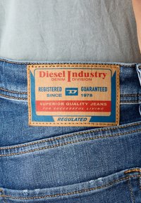 Denim-farkut, joissa on ruskea nahkalappu, jossa on punaisia ja sinisiä tekstejä. Lappu ilmoittaa "Diesel Industry", "Superior Quality Jeans" ja siinä on ompeleiden yksityiskohtia.