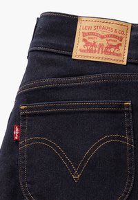 Jeans de mezclilla oscura con un parche de cuero marrón que presenta el logo de "Levi Strauss & Co.", costuras amarillas y una pequeña etiqueta roja en el bolsillo.