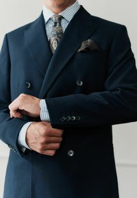 Tumesinine kahekupeeline ülikond, villane materjal, millel on suured nööbid, kontrastse helesinise triibulise särk ja mustriga lips koos taskurätikuga.