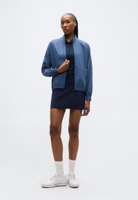 Γυναίκα που στέκεται, φοράει μπλε ζακέτα με φερμουάρ πάνω από μπλε φόρεμα, λευκές κάλτσες και λευκά αθλητικά παπούτσια, μπροστά σε απλό ανοιχτό φόντο.