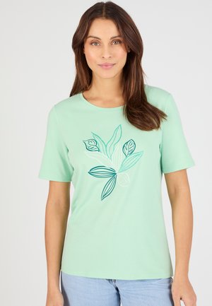 Femme portant une chemise à manches courtes vert clair avec un motif abstrait de feuilles en bleu sarcelle et blanc, associée à un jean bleu clair.