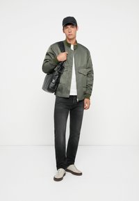 Green bomber jacket with a high collar, paired with dark jeans and beige sneakers. Black bag slung over shoulder, white t-shirt underneath.
