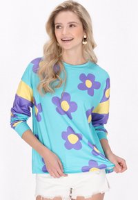 Camisa de manga larga en tejido turquesa adornada con grandes flores moradas y centros amarillos. Presenta mangas a rayas amarillas y moradas.