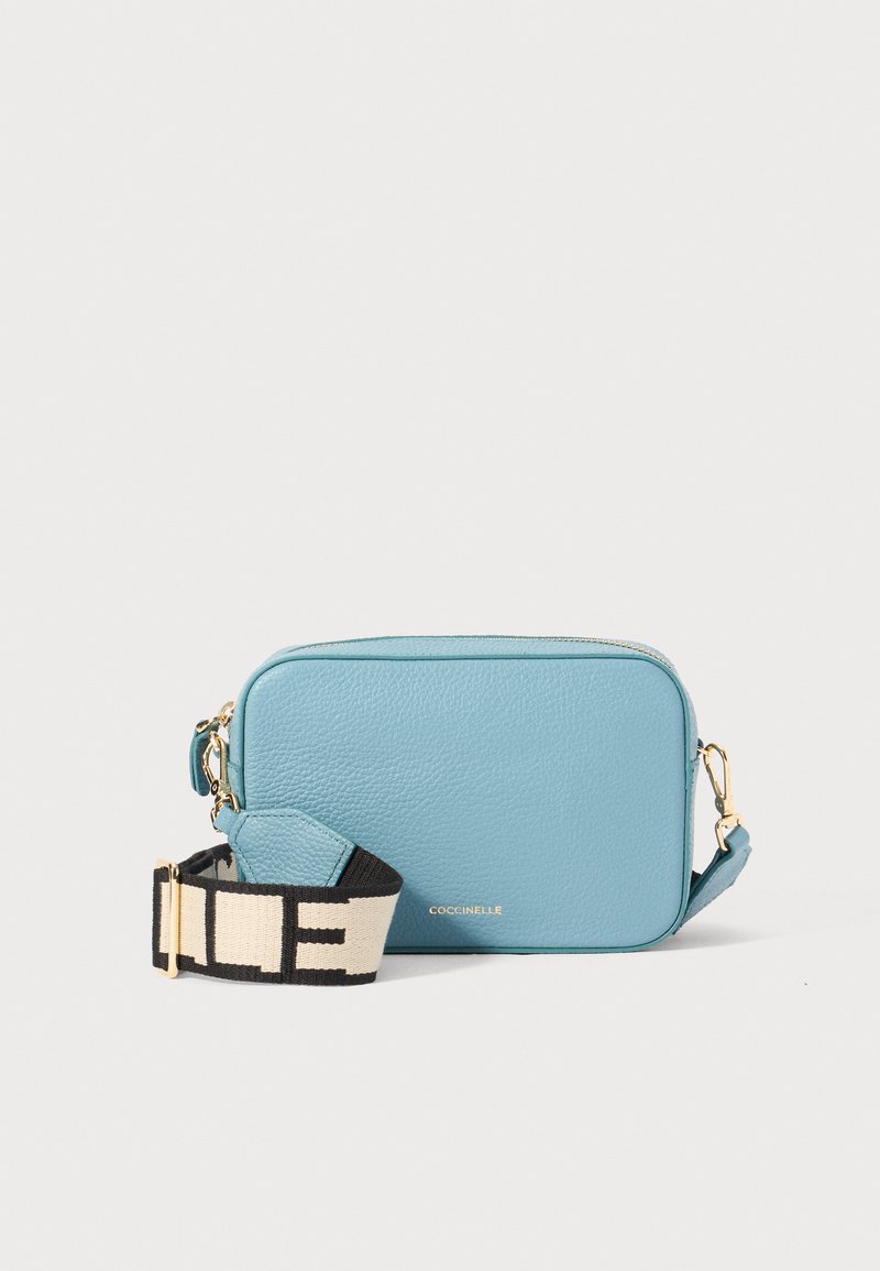 Bolsa transversal de couro azul com uma textura suave, forma retangular e uma alça padrão em preto e creme. Acessórios em dourado visíveis.