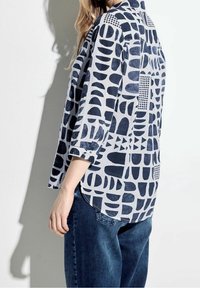 Chemise bleu et blanche à motifs, présentant des formes abstraites et des lignes, avec des manches longues, un col et un ourlet arrondi. Associée à un jean en denim.