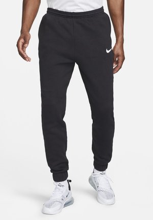 Homme portant un pantalon de jogging noir Nike avec des poignets élastiques et des baskets blanches Nike Air 270 sur fond blanc.