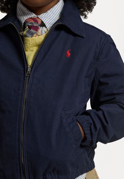 Marineblaue Zip-Jacke mit Kragen, elastischen Bündchen und einem roten gestickten Logo. Über einem gelben Sweater und einer gemusterten Krawatte getragen.