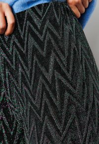 Tissu noir avec un motif en zigzag dans des tons verts et violets scintillants, créant un effet visuel texturé. Vue en gros plan, main tenant le bord.