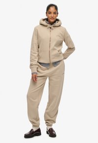 Felpa beige con zip frontale, cappuccio con cordino e tasche anteriori, abbinata a pantaloni tapered coordinati, presentata con una camicia a strisce stratificata.