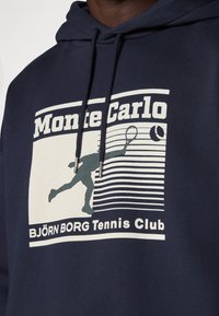 Σκούρο μπλέ φούτερ φτιαγμένο από βαμβάκι με γραφική εκτύπωση που απεικονίζει έναν παίκτη τένις, κείμενο "Monte Carlo" και "BJÖRN BORG Tennis Club."