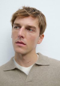 Beige gebreide polo met een kraag en textuurstof, gelaagd over een witte crewneck shirt, tegen een lichte achtergrond.