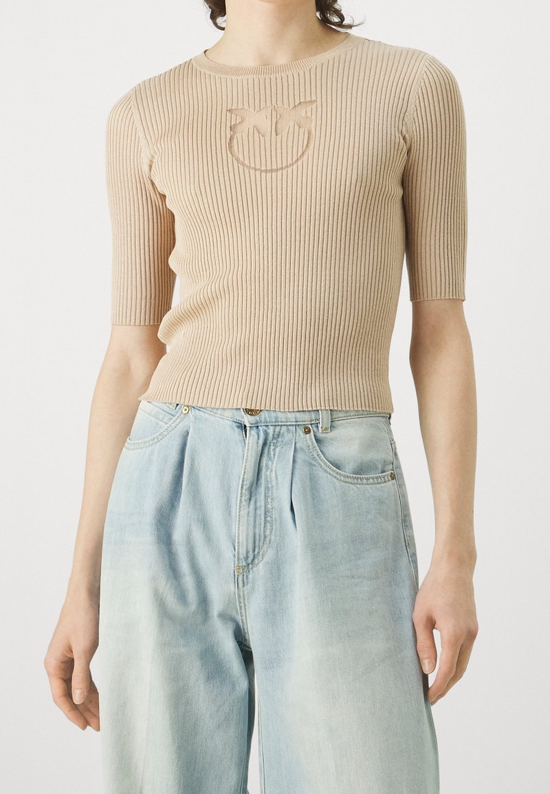 Haut beige à manches courtes en côte avec un logo subtilement embossé, associé à un jean clair taille haute à coupe décontractée porté par une personne debout.