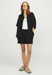 Camisa negra de botones con un corte relajado y mangas cortas, combinada con pantalones cortos negros. El modelo lleva una blusa blanca y zapatillas sin cordones beige.