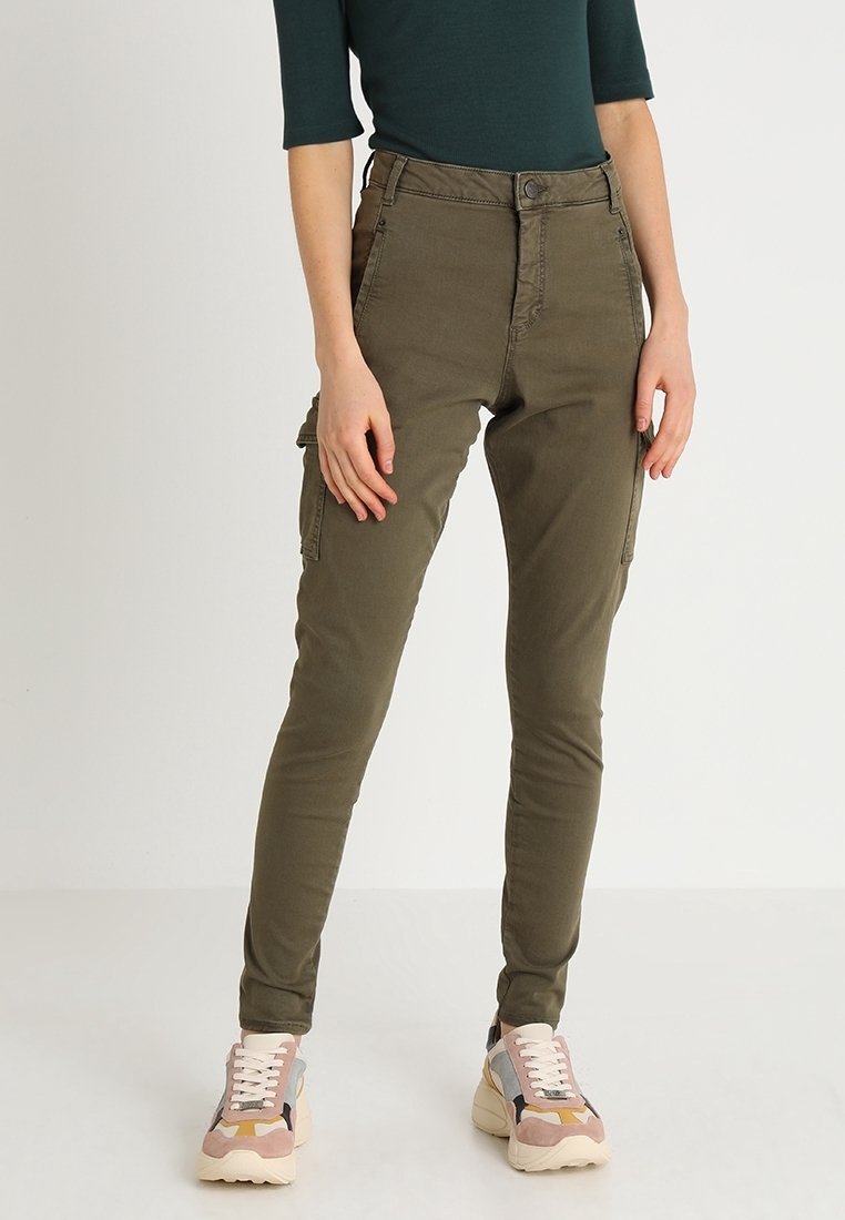 Pantalon cargo vert olive, avec une taille haute, une coupe slim, des poches avant et latérales, en tissu lisse et résistant. Associé à des baskets multicolores.