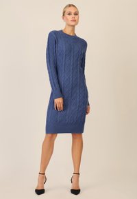 Abito blu lavorato a maglia con motivo a treccia, maniche lunghe e bordo a costine. Indossato con scarpe con tacco alto nere.
