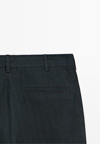 Ciemnoszare spodnie w prążki, widok z tyłu ukazujący pojedynczą kieszeń z patką i szlufki na pasek na białym tle.