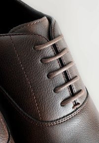 Chaussure en cuir brun avec une surface texturée, dotée d'un design traditionnel à lacets et de coutures renforcées le long des bords.