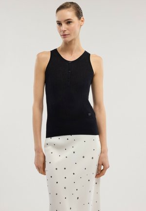 Frau trägt ein ärmelloses schwarzes, geknöpftes Oberteil und einen weißen Rock mit verstreuten schwarzen Punkten, steht vor einem schlichten hellen Hintergrund.