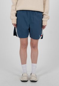 Person trägt marineblaue Sportshorts mit weißem und rotem Logo, cremefarbenen Pullover, weiße Socken und cremefarbene Loafers und steht auf einem grauen Boden.