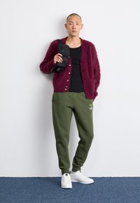 Burgundin värinen pörröinen neuletakki, musta toppi, vihreät verkkarit Nike-logolla, valkoiset lenkkarit, pienen mustan laukun pitäminen, seisoo valkoista seinää vasten.