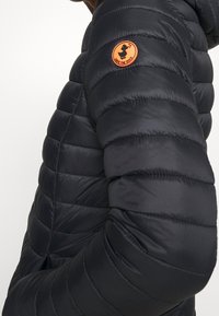 Sort quiltet pufferjakke med hætte, tæt på armen med en orange rund lap, der har et ande logo og teksten "SAVE THE DUCK".