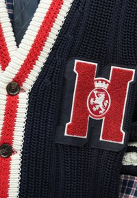 Σκούρο μπλε καρδιογκαντίνα με κόκκινες και λευκές ρίγες, με υφή και κεντημένο έμβλημα σε σχήμα 'H' και κουμπιά. Πλεκτό ύφασμα και στρωματοποιημένος γιακάς με καρό μοτίβο.