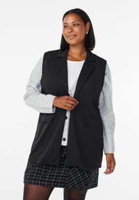 Blazer senza maniche nero con un design su misura, caratterizzato da due tasche frontali, indossato sopra una maglietta a maniche lunghe grigia chiara e una gonna con fantasia.