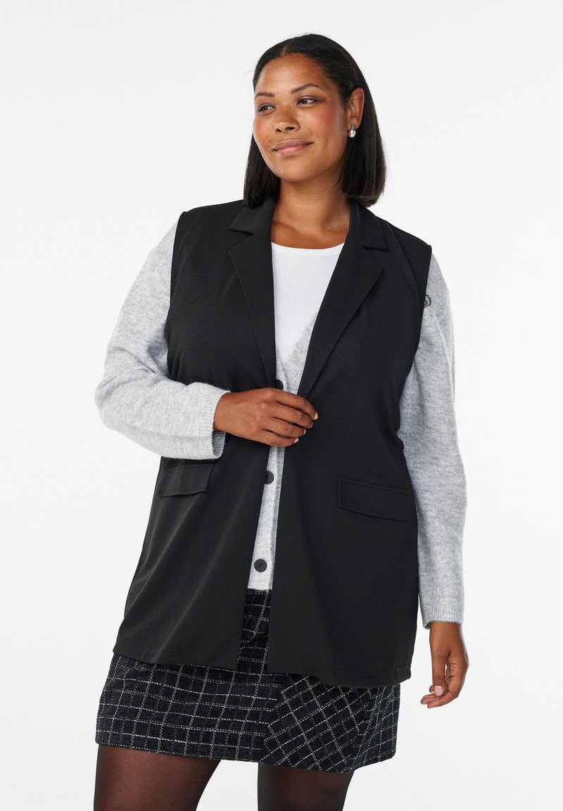 Blazer senza maniche nero con un design su misura, caratterizzato da due tasche frontali, indossato sopra una maglietta a maniche lunghe grigia chiara e una gonna con fantasia.