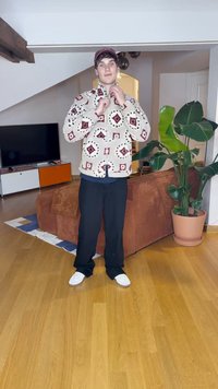 Bela flisasta jakna z rdečimi in črnimi diamantnimi vzorci, črne hlače in bele tenisice. Notranji ambient z rjavim kavčem in rastlino.