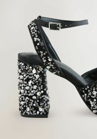 Black high-heeled sandals with a chunky heel covered in sequins. Features a thin ankle strap and soft material on the upper portion.