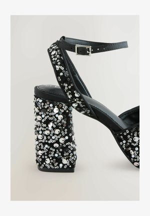 Sandale negre cu toc înalt, cu un toc gros acoperit de paiete. Dispun de o bră?ară sub?ire pentru gleznă și un material moale în partea superioară.