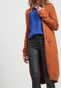 Orange stickad lång kofta med ribbade ärmar och fickor, bärs över en blå skjorta med knappar och svarta byxor. Texturerad, mjuk tyg.