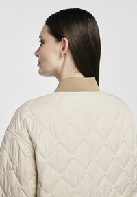 Beżowa pikowana kurtka z diamentowym wzorem, prążkowany beżowy kołnierz oraz gładka faktura. Widok z tyłu pokazuje przeszycia wzdłuż środka.