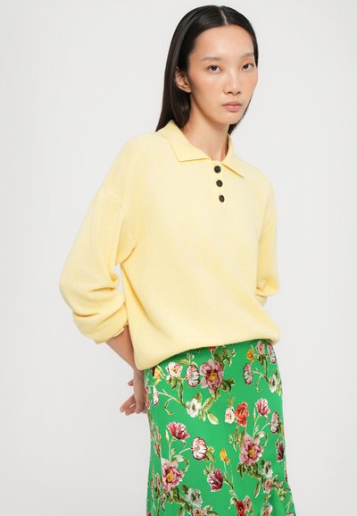 Maglione giallo con colletto e tre bottoni neri, abbinato a una gonna verde con stampa floreale che presenta fiori rossi e rosa. Tessuto morbido.