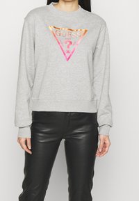 Šedá mikina s trojúhelníkovým grafickým logem v růžové a oranžové barvě. Dlouhé rukávy a kulatý výstřih. Nosí se s černými koženými kalhotami.