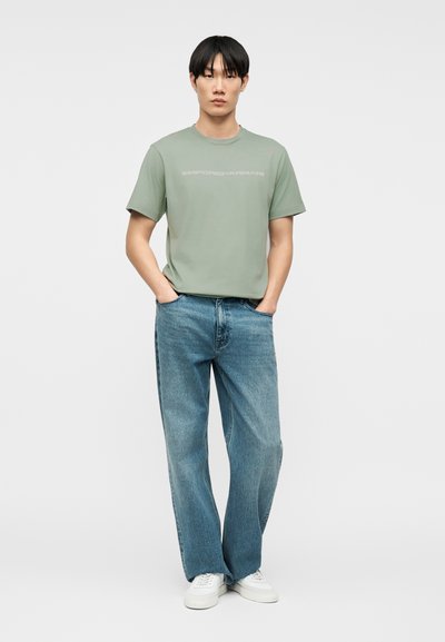 Νέος άνδρας που στέκεται με τα χέρια στις τσέπες, φοράει μια μπλούζα Emporio Armani σε πράσινη απόχρωση φασκόμηλου, άνετα μπλε τζιν και λευκά αθλητικά παπούτσια.