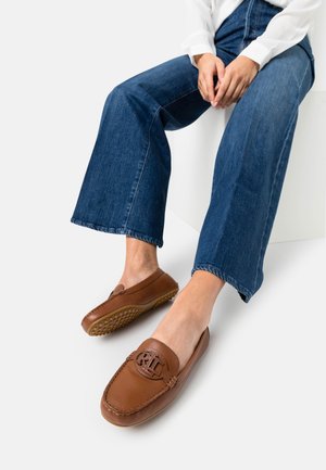 Mocassins en cuir brun avec une semelle en caoutchouc texturé et un logo embossé. Associés avec un jean bleu évasé, mettant en valeur le design décontracté de la chaussures.