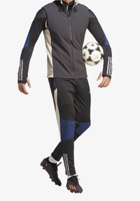 Chaqueta deportiva negra con acentos en azul y beige, con cuello de cremallera, combinada con pantalones negros a juego que presentan secciones azules. Botas de fútbol negras.