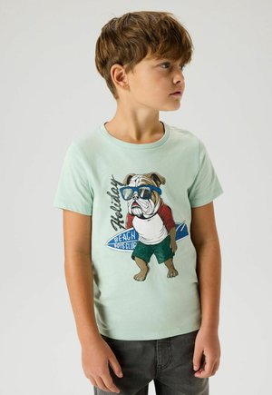 Chłopiec patrzący w prawo, ubrany w jasnooliwkowy t-shirt z grafiką buldoga w okularach przeciwsłonecznych, czerwoną i białą koszulę, zielone szorty oraz deskę surfingową.